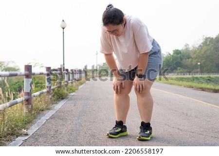 Wide shot portrait of overweight Asian woman wearing eyeglasses, standing outdoor, bending down, resting after running, being tired, suffering from knee and joint injury. Sport and obesity concept. Royalty-Free Stock Photo #2206558197