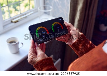 Senior Woman With Smart Meter Trying To Keep Warm By Radiator During Cost Of Living Energy Crisis Royalty-Free Stock Photo #2206567935