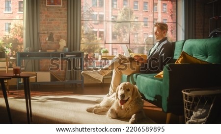 Handsome Young Man Using Laptop, Working from Home Living Room with Big Windows. Male Sitting on Couch, Using Computer, Golden Retriever Dog Sitting Next to Him and Wanting to Play or Go For a Walk. Royalty-Free Stock Photo #2206589895