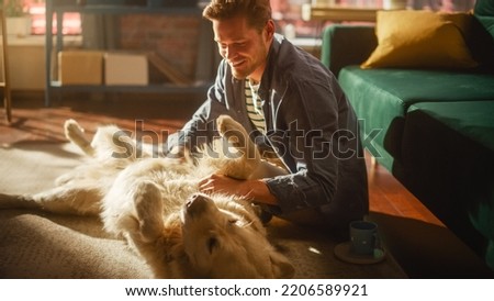 Successful Young Adult Man Playing with His Dog at Home, Active Golden Retriever. Man Sitting on a Floor Teasing, Petting and Scratching an Excited Dog, Having Fun in the Loft Living Room Space. Royalty-Free Stock Photo #2206589921