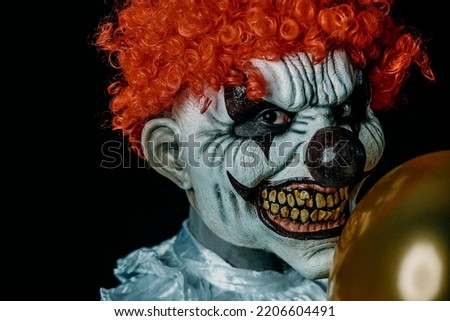 closeup of a creepy evil clown with red hair, staring at the observer, holding a golden balloon in front of him, against a black background