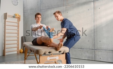 Young Male Athlete Undergoing Physiotherapy, Professional Sport Masseur Helping with Foot Exercise with a Rubber Band. Musculoskeletal Pain Therapy and Rehabilitation in Modern Clinic Concept. Royalty-Free Stock Photo #2206610153