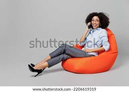 Full body fun young employee business corporate lawyer woman of African American ethnicity in classic shirt work in office sit in bag chair talk speak on mobile cell phone isolated on grey background