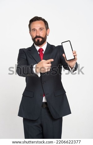 Handsome mature businessman in black suit pointing at smartphone mock up white screen looking at camera isolated on white background. Beard Businessman in suit. App Product placement.