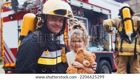 Firefighter hug rescued little girl with teddy bear. Frightened child rejoices in rescue. At background firefighters after extinguishing fire next to fire truck. Concept of saving lives, fire safety