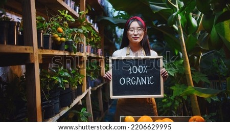 Close up of charming asian woman in glasses standing in greenhouse behind box with fruits, holding wooden sign organic, showing to camera and smiling. Hobby farming, agriculture concept.