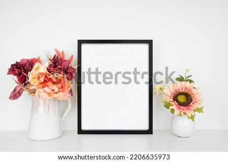 Mock up black picture frame with pink hue autumn flowers on a wood shelf against a white wall. Fall concept. Copy space.