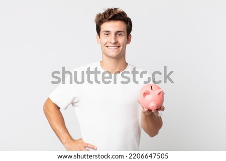 young handsome man smiling happily with a hand on hip and confident and holding a piggy bank