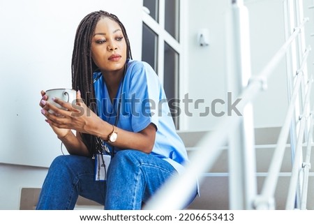 A woman wearing blue scrubs walks through the halls of a hospital or clinic while holding a coffee cup in one hand. Portrait of black nurse drinking coffee. 