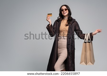 smiling young woman in stylish sunglasses holding shopping bags and credit card isolated on grey Royalty-Free Stock Photo #2206655897