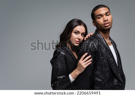young woman in leather jacket hugging stylish african american man isolated on grey Royalty-Free Stock Photo #2206659491