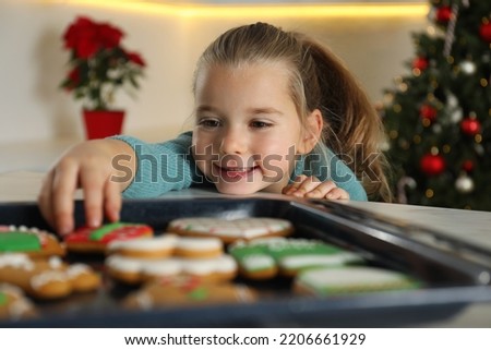 Cute little girl taking fresh Christmas gingerbread cookie from baking sheet at table indoors