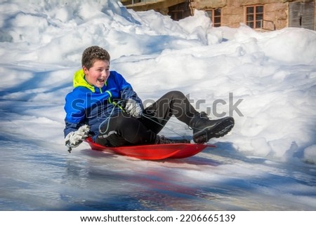 On a bright sunny day in winter, a boy slides down a hill on a plastic sled on his stomach. He is joyful.