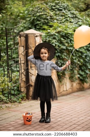 Little cute girl in witch costume holding balloon, Nearby stand jack-o-lantern pumpkin bucket with candies and sweets. Kid trick or treating in Halloween holiday