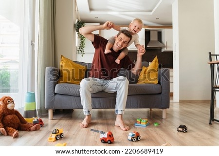 Happy father carrying his laughing little blond son on neck and sitting on couch in living-room surrounded with toys, spending time together on weekend. Young dad playing with his toddler boy Royalty-Free Stock Photo #2206689419