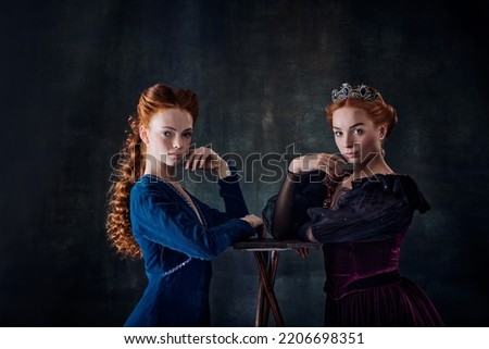 Queens. Two beautiful redhaired women, princess looking at camera over dark vintage background. Royal family. Concept of modernity and renaissance, baroque style, beauty, history