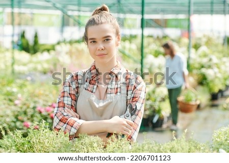 Photo realistic digital collage of young female gardener in casualwear and apron standing against large flowerbeds and colleague in hothouse