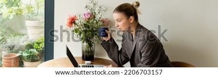Woman in jacket holding cup and using laptop near flowers at home, banner