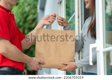 Happy smiling Asian woman receives paper bag of food from courier in front  house. Delivery man in red uniforms deliver express. online shopping, paper containers, takeaway, postman, delivery service Royalty-Free Stock Photo #2206705019