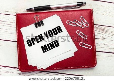 On a light wooden background, a burgundy pen and notebook. On the notebook has white paper clips and white paper with the text OPEN YOUR MIND. Business concept