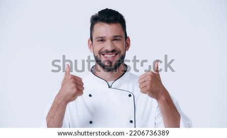 happy young chef showing thumbs up and looking at camera isolated on white