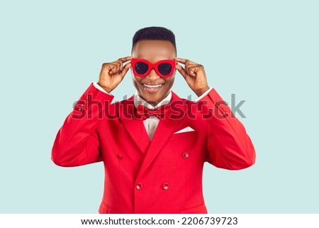 Funny young African American man millennial in red suit for work host of entertainment event or party puts on sunglasses over eyes and looks at camera smiling stands on light studio background