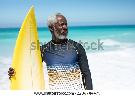 Front view of a senior African American man on a beach in the sun, holding a surfboard and looking away, with blue sky and sea in the background