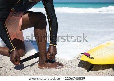 Low section side view of a senior African American man on a beach in the sun, kneeling and attaching the leash of a surfboard to his leg, with blue sky and sea in the background Royalty-Free Stock Photo #2206745839