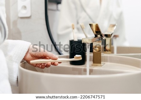 Concept of oral and dental care at home. Cropped shot of woman holding bamboo toothbrush with toothpaste over sink. Female in bathrobe standing in bathroom in morning Royalty-Free Stock Photo #2206747571