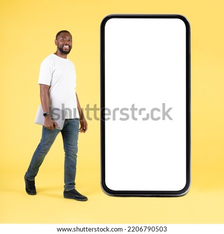 Happy Black Guy Near Large Smartphone Blank Screen Posing Holding Laptop Walking On Yellow Background In Studio. Mobile Application Advertisement. Square, Full Length Shot