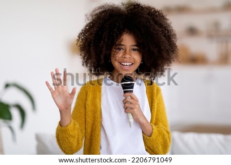 Cute preteen black girl with bushy hair holding microphone and waving at camera, talented child singing karaoke at home, recording songs for contest. Children's lifestyle concept
