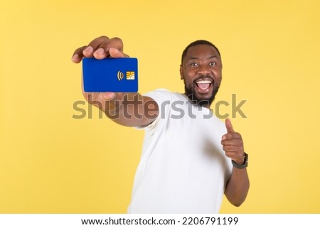 Happy African American Male Showing Blue Credit Card To Camera Approving His Bank Gesturing Thumbs Up Smiling To Camera Standing On Yellow Background In Studio. Financial Offer. Selective Focus