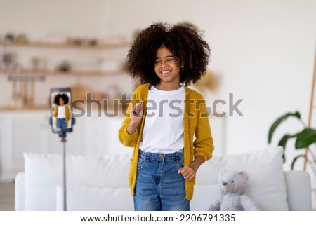 Beautiful preteen black girl with cute hair recording video for her friends while having fun at home, standing in front of smartphone set on tripod, dancing and smiling, copy space