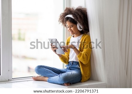 Cheerful preteen black girl with bushy hair using modern digital tablet and wireless headset, sitting on windowsill at home, playing mobile game or checking newest app, touching pad screen, copy space