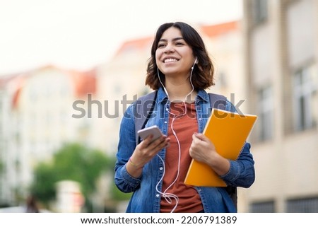 Cheerful young arab student female listening music with smartphone and earphones outdoors, smiling middle eastern woman carrying workbooks and backpack while walking on city street, copy space Royalty-Free Stock Photo #2206791389