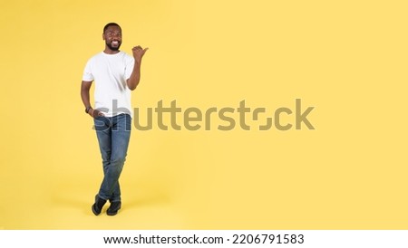 Great Offer. Cheerful African American Guy Pointing Thumb Finger Aside At Copy Space Standing Over Yellow Studio Background. Look There Concept. Full Length, Panorama