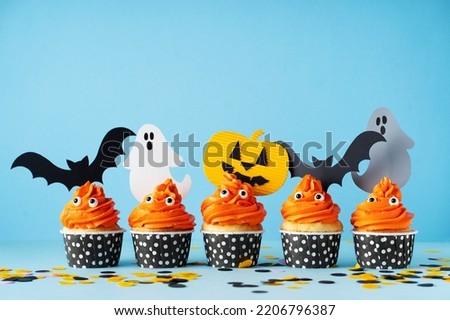 Monster orange funny cupcakes with eyes and ghost and bat on blue background, copy space