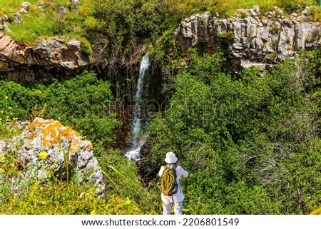 Woman with backpack in white hat photographs picturesque landscape. Picturesque waterfall in the basalt gorge. Jewish Easter in Israel. Flowering green slopes of the Golan Heights. 
 Royalty-Free Stock Photo #2206801549
