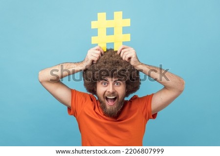 Portrait of man with Afro hairstyle holding yellow hashtag sign board on head, dreaming about target tagging posts on his website, smm manager. Indoor studio shot isolated on blue background. Royalty-Free Stock Photo #2206807999