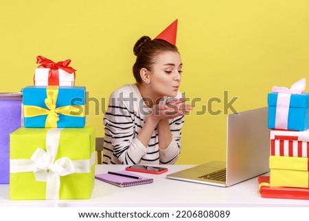 Charming woman in party cone sending air kisses to laptop display, sitting at workplace, receive congratulations from friends by video call. Indoor studio studio shot isolated on yellow background.