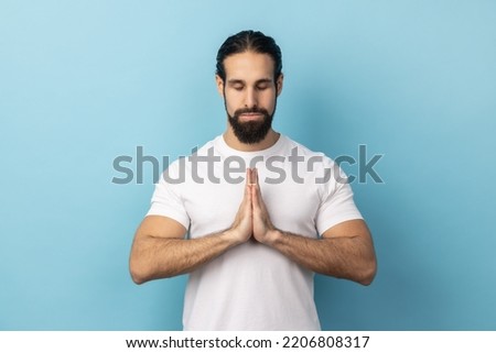 Man with beard wearing white T-shirt concentrating his mind, keeping hands namaste gesture, meditating, yoga exercise breath technique reduce stress. Indoor studio shot isolated on blue background.