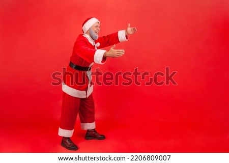 Full length profile shot of elderly man with gray beard wearing santa claus costume reaching hands out, stretching arms to hug someone. Indoor studio shot isolated on red background.