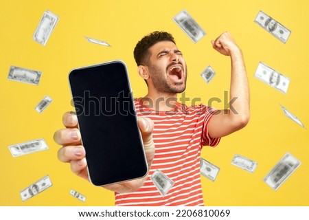 Money rain, winner and rich. Portrait of rejoicing happy bearded man wearing red T-shirt standing with smart phone in hands, showing blank screen. Indoor studio shot isolated on yellow background. Royalty-Free Stock Photo #2206810069