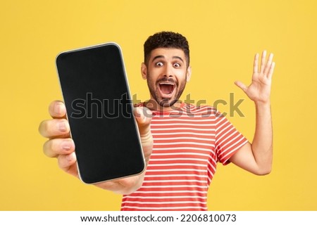 Amazed cheerful man with beard wearing red T-shirt showing mobile phone with blank display, expressing happiness, raised arm and screaming happily. Indoor studio shot isolated on yellow background. Royalty-Free Stock Photo #2206810073