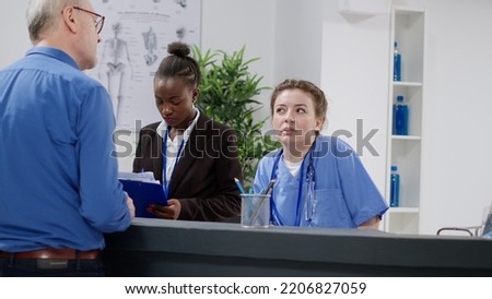 Diverse medical team working at hospital reception desk, helping patients with checkup appointments and report papers. Nurse and receptionist sitting at registration counter in facility lobby.