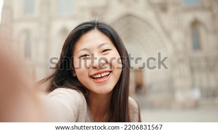 Beautiful Asian girl sincerely smiling while taking selfie against the backdrop of old architecture. Japanese woman looking happy exploring new city