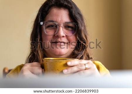 Close-up of a woman holding a coffee in her hands while in a work meeting on her computer at home. Yellow