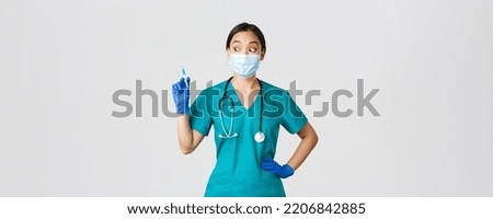 Covid-19, coronavirus disease, healthcare workers concept. Excited asian female nurse in medical mask, rubber gloves and scrubs, looking amazed at syringe with vaccine, white background