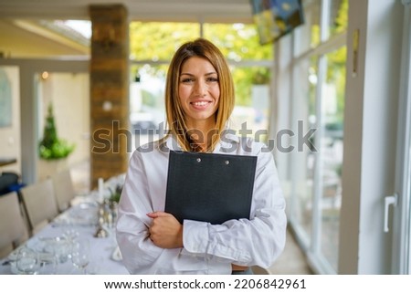 One woman young adult caucasian standing and smiling in the restaurant or hotel with paperwork in her hands and waiting clients Entrepreneur manager business modern lifestyle concept