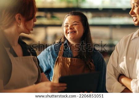 Retail worker with Down syndrome smiling happily while standing in a staff meeting in a grocery store. Group of diverse women working together in an all-female small business. Royalty-Free Stock Photo #2206851051